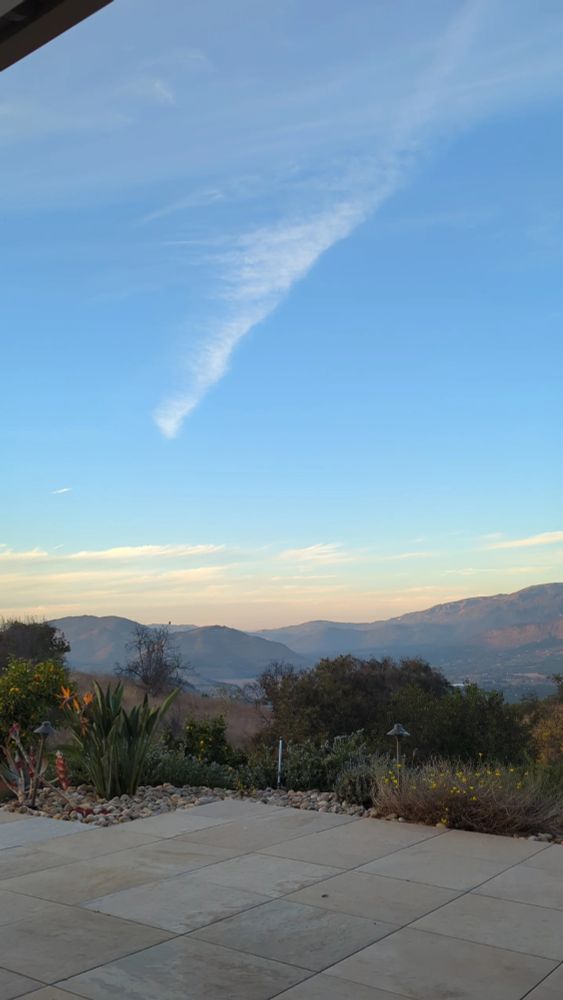 Photo of an outdoor patio overlooking chaparral, orange trees, and blue-green mountain range in the distance. It's early evening and the sky is a cornflower blue, with a single brush stroke of cirrus cloud dragging the eye upwards, while low crowds of hazy pink-orange clouds nestle against the horizon, skimming the tips of the distant mountains.