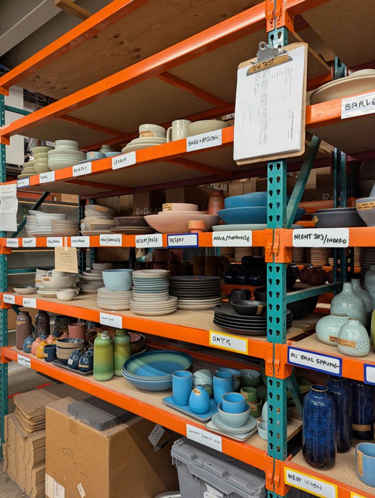 Photo of a quality assurance shelf at Heath Ceramics Factory. Various plates, saucers, cups and vases sit grouped by color, with ideal color samples to ensure that glazing looks correct.