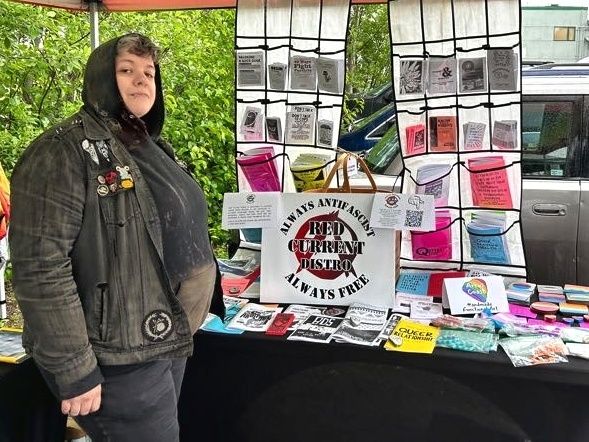A person dressed in black with a hood on stands next to a table display of zines, pamphlets, & other printed material. There is a sign for Red Current Distro which reads, "Always antifascist, always free."