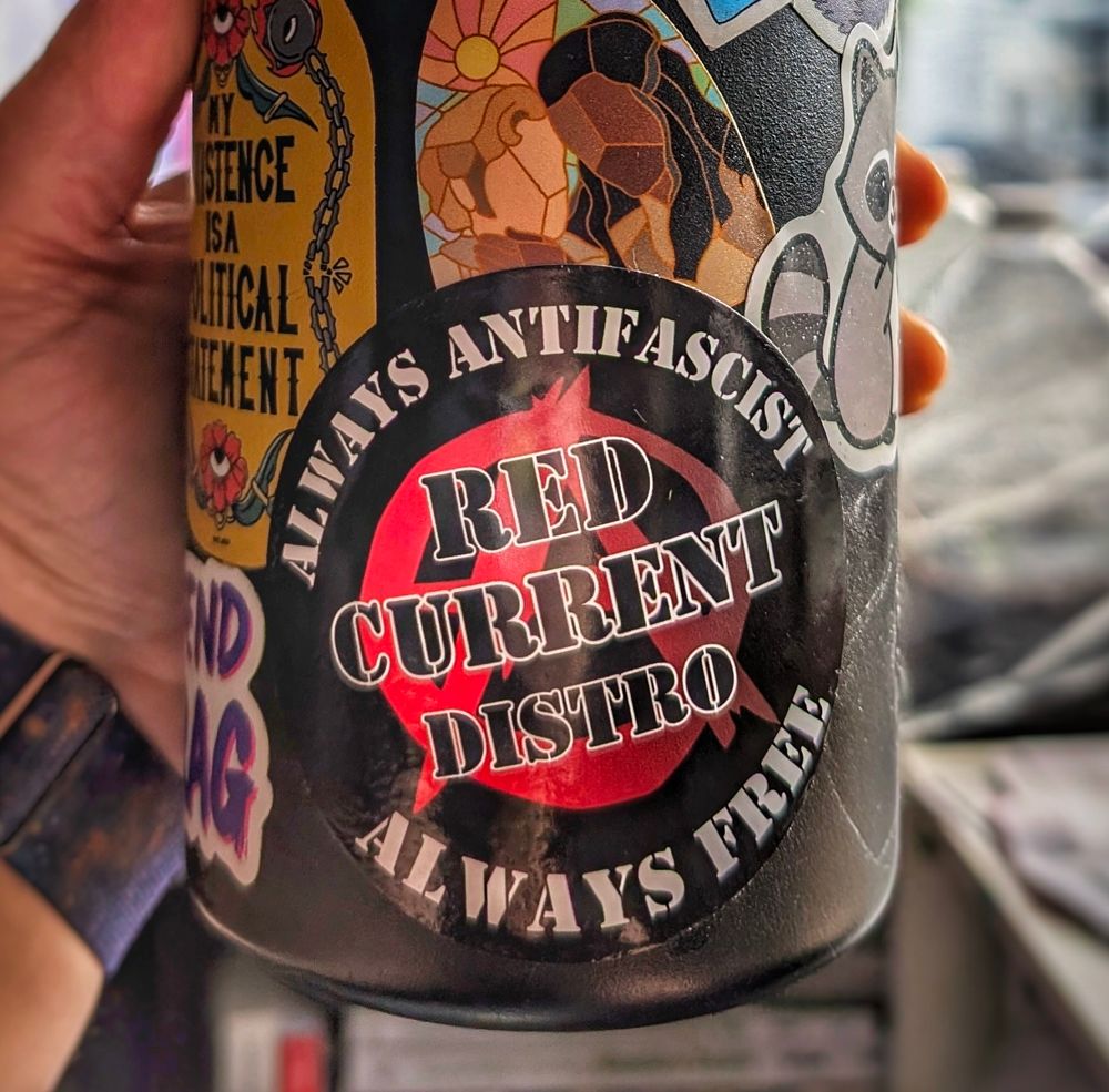 The bottom of a black water bottle covered in stickers. The focus at the center of the picture is a round black sticker with the words "Always antifascist" on top & "always free" on bottom. In the center is a red anarchist a with the name "Red Current Distro" over it.