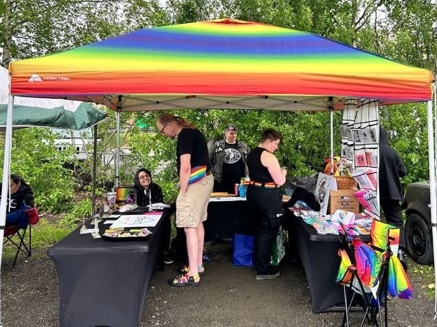 A rainbow pop up tent at an outdoor event with 3 tables covered in black cloth displaying items & information. Two people browse the tables & two people engage with them about the items they're presenting.