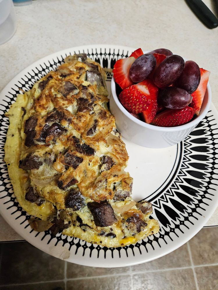 A black and white plate with a steak and Caramelized Onions omelet on the left side, and a white bowl with strawberries and red grapes on the right
