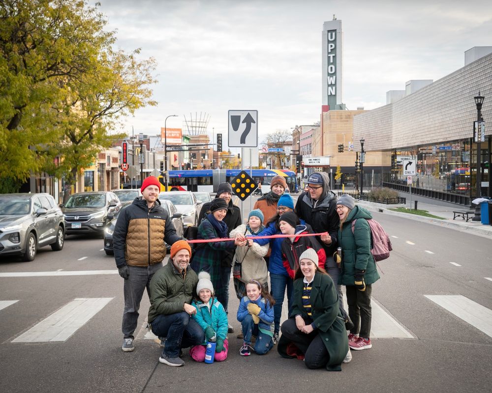 A group of people pose in the middle of Hennepin Ave wearing winter coats. In the center, a make-shift ribbon cutting is taking place 