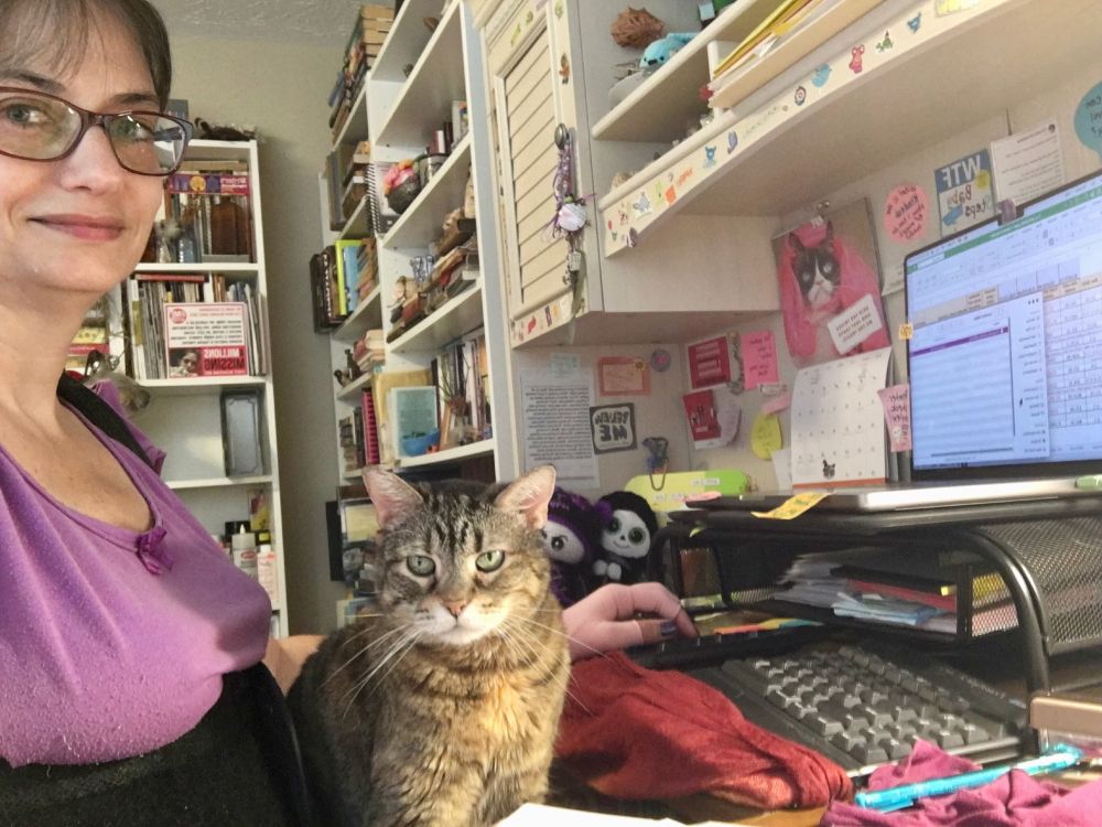 A tabby cat staring at the camera sits on the lap of a white female wearing a purple shirt, purple glasses, and black back brace. She is sitting at her computer and surrounded by a very messy desk.