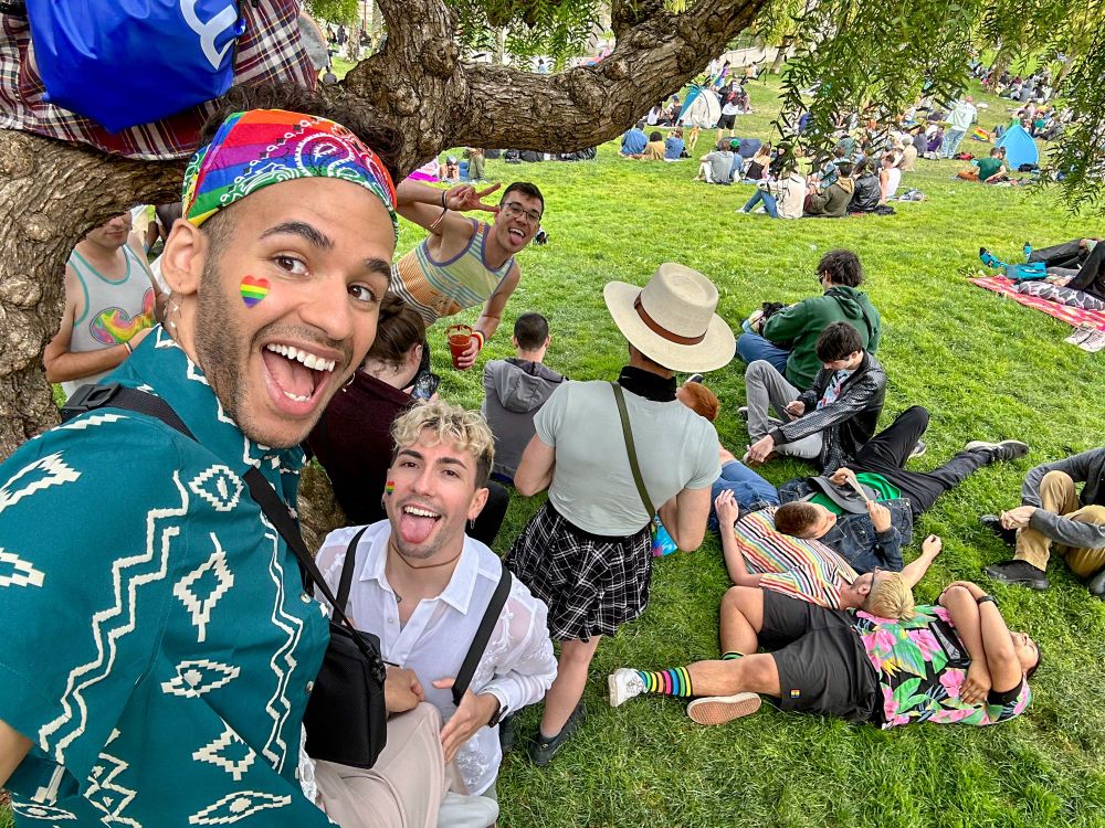 Mikah Sargent and his partner, Sebastian, smile and stick their tongue out, respectively. They are with a large group of individuals resting on the lawn and a tree at Dolores Park.
