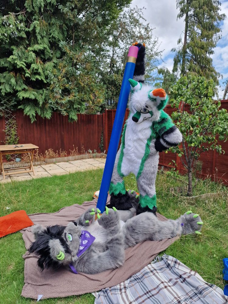 A fursuiter stood over another holding a giant pencil against his chest.