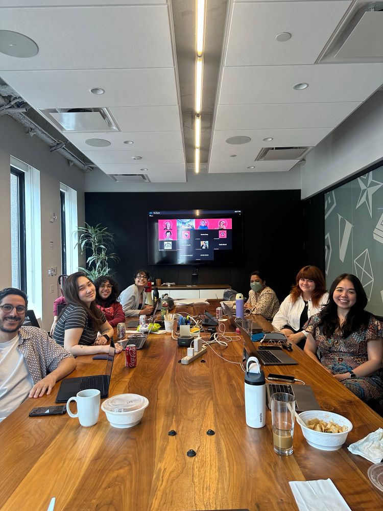 People seated at a meeting room conference table with people on a zoom meeting in the back.