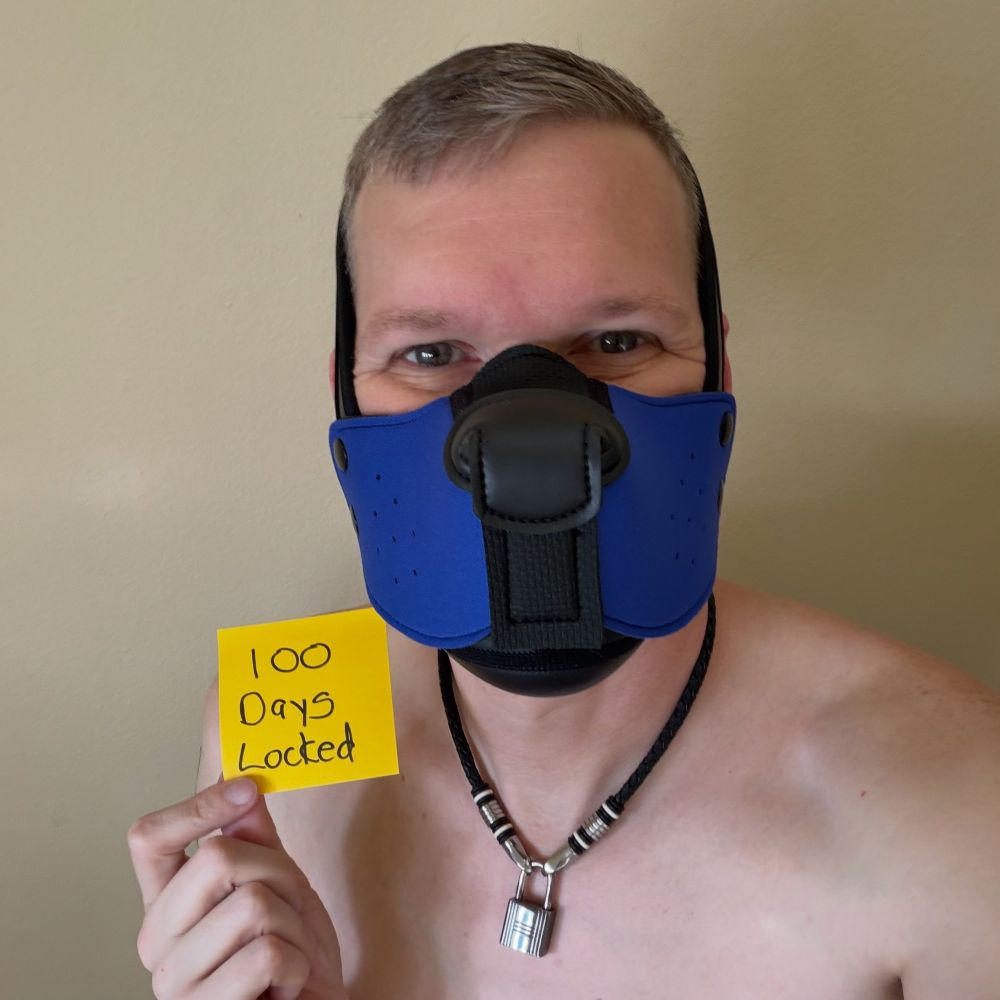 A human pup in a blue half hood holds up a post it that says “100 days locked”