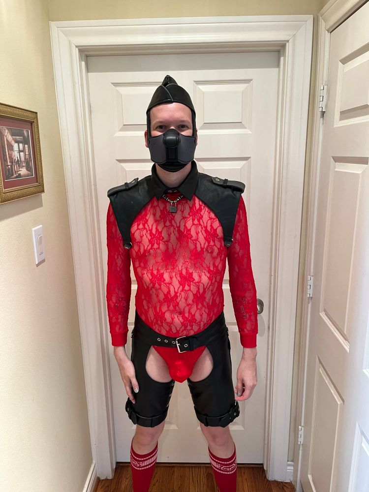 A human pup wearing a red lace unitard with black leather chaps, a black brigadier harness, and garrison cap. 