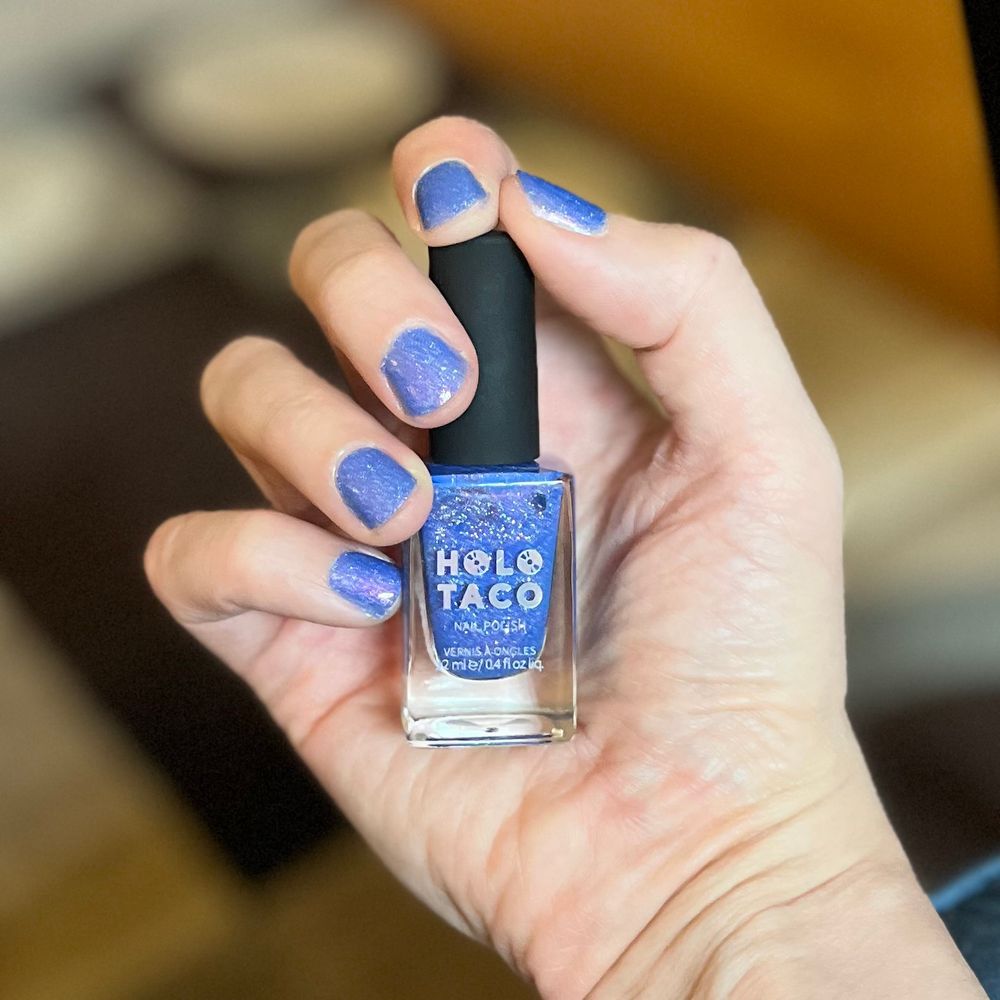 A hand holds a bottle of light blue glittery Holo Taco nails polish. 