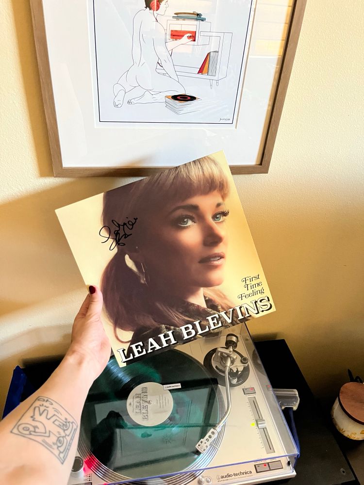 An extended arm holds a vinyl copy of First Time Feeling by Leah Blevins over a record player. 

Artwork on the wall features a a naked man sitting in front of a record player. 