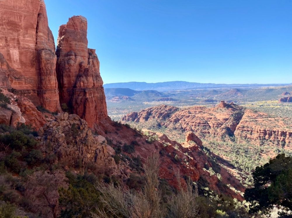 Cathedral Rock, Coconino National Forest. Tall, sienna, tan, and brown sandstone butte formations that make up Cathedral Rock stand in the far left side. At there base is dark green foliage and scrub plants in the foreground. Below and in the middle and right side are the lower tan, sunlit mesas and scattered green foliage and the Oak Creek canyon floor. In the background are the blue appearing buttes and mesas under a clear blue early afternoon sky in November.