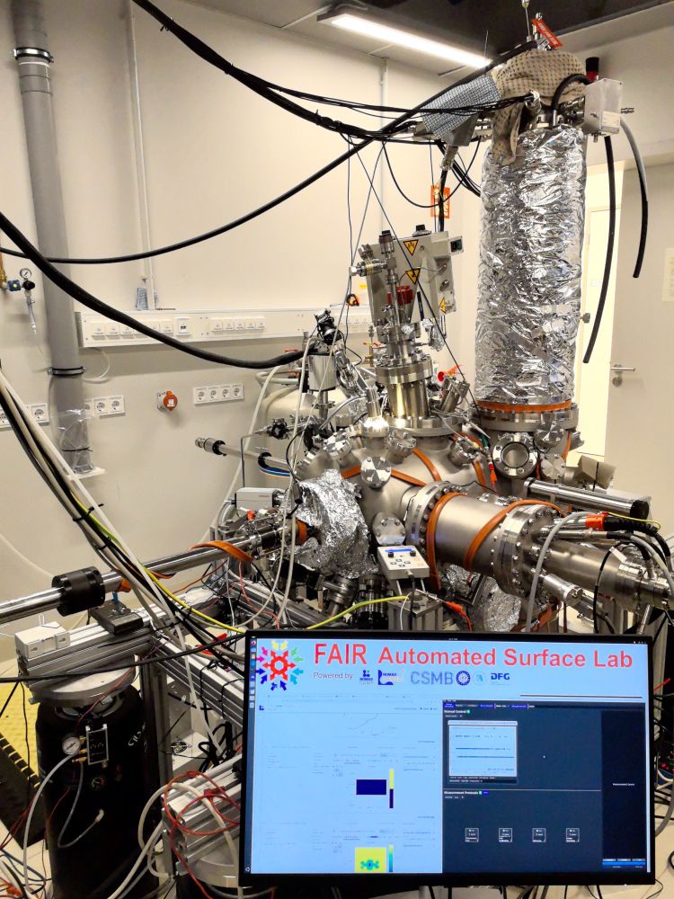A ultra-high vacuum lab featuring a shiny metal chamber with a photoemission electron microscope coupled to a 4 K scanning probe microscopy. The photograph also depicts a monitor which reads "FAIR Automated Surface Lab" and two panels, the lhs panel contains scanning probe spectroscopy and scanning tunneling spectroscopy HDF5 datasets being uploaded in the local lab NOMAD Oasis. The rhs panel is running a temperature measurement protocol for the sample.  