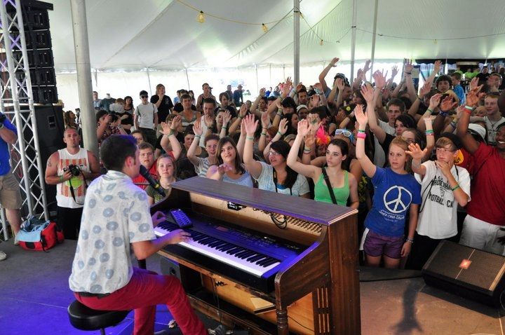 We see a young man (Tyler Joseph). The picture was taken from the stage, facing the crowd. Tyler is sitting and playing his brown saloon-style piano and singing.
He's wearing red long pants and a white short sleeve button-up shirt with decent light blue patterns.
The people in the crowd are looking at him, and almost all of them hold their right arm up.
It's probably warm, as they all wear light clothing and the venue is a large white tent.