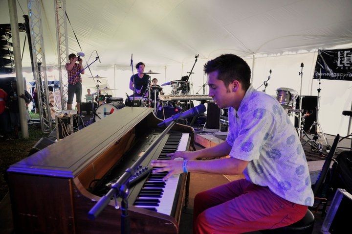 We see a young man (Tyler Joseph) close up from the left side of the stage. He's playing his brown saloon-style piano and singing.
He's wearing red long pants and a white short sleeve button-up shirt with decent light blue patterns. In the background, we can see the bands drummer (Josh Dun) playing and their friend Mark (known as reelbearmedia) who is taking photos of the concert.
 The venue is a large white tent. There is no crowd visible in this photo.