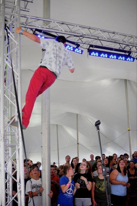 While performing, a young man (Tyler Joseph) has climbed up the stages side scaffolding and is singing to a crowd while only holding on to it with one hand.
He's wearing red long pants and a white short sleeve button-up shirt with decent light blue patterns.
The people in the crowd are looking up to him with amazed and smiling face impressions.
