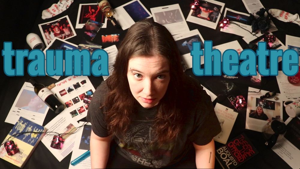 Laura sits on the floor surrounded by computer printouts, books, empty bottles and glasses, and string lights. Blue text across the image reads "trauma theatre".
