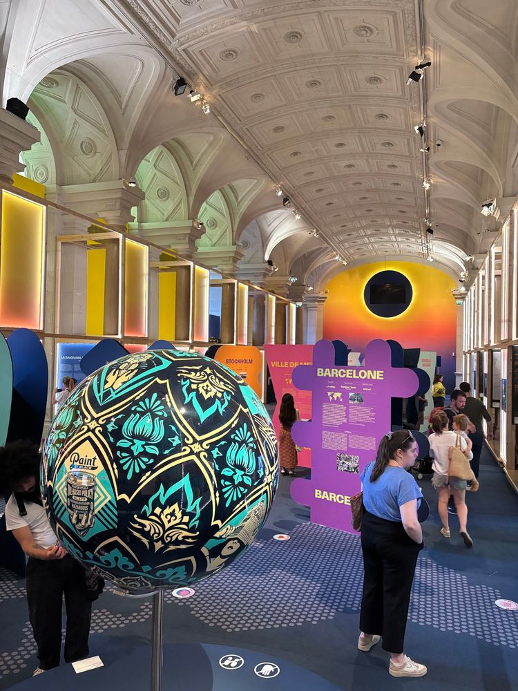 A wide angle shot of the exhibition hall. It’s all purples, oranges and yellows, with a teal and yellow painted sphere in the foregound. Puzzle-piece shaped panels fill the hall, each one highlighting a global city and a climate/human-focused project it has successfully launched. 