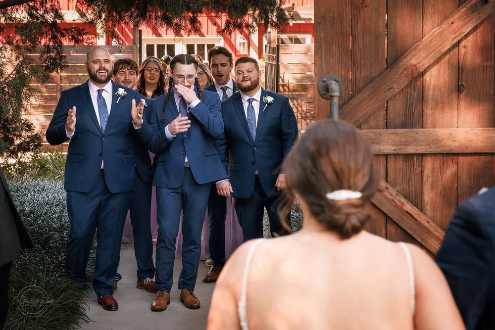 5 groomsman in matching suits standing next to one another as a door opens to reveal a first look at the bride in her dress and the groom with her, everybody else is excited and i am genuinely crying—the bridesmaids trail us but only bc they already seen the bride i promise