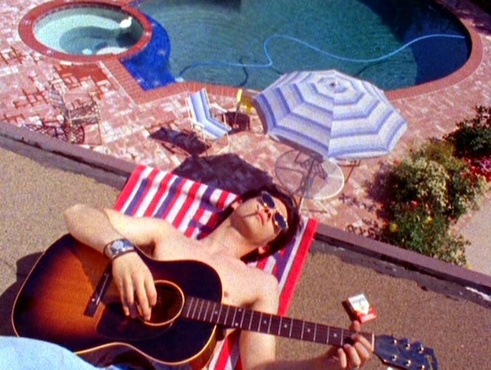 a man lays on a towel playing guitar, in the background you see a swimming pool