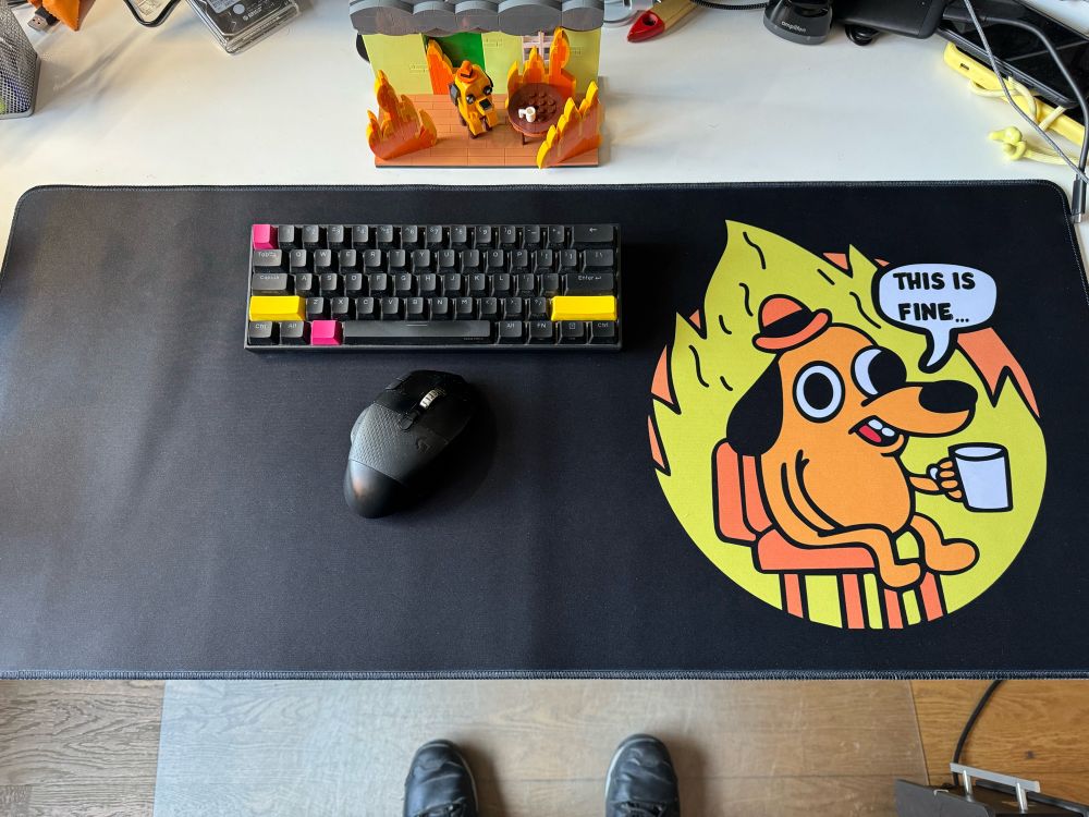 Image shows a huge mouse pad with “This is fine” dog meme image on it, also shows a key board an mouse on top of the pad and at the top of it there is a Lego version of the meme