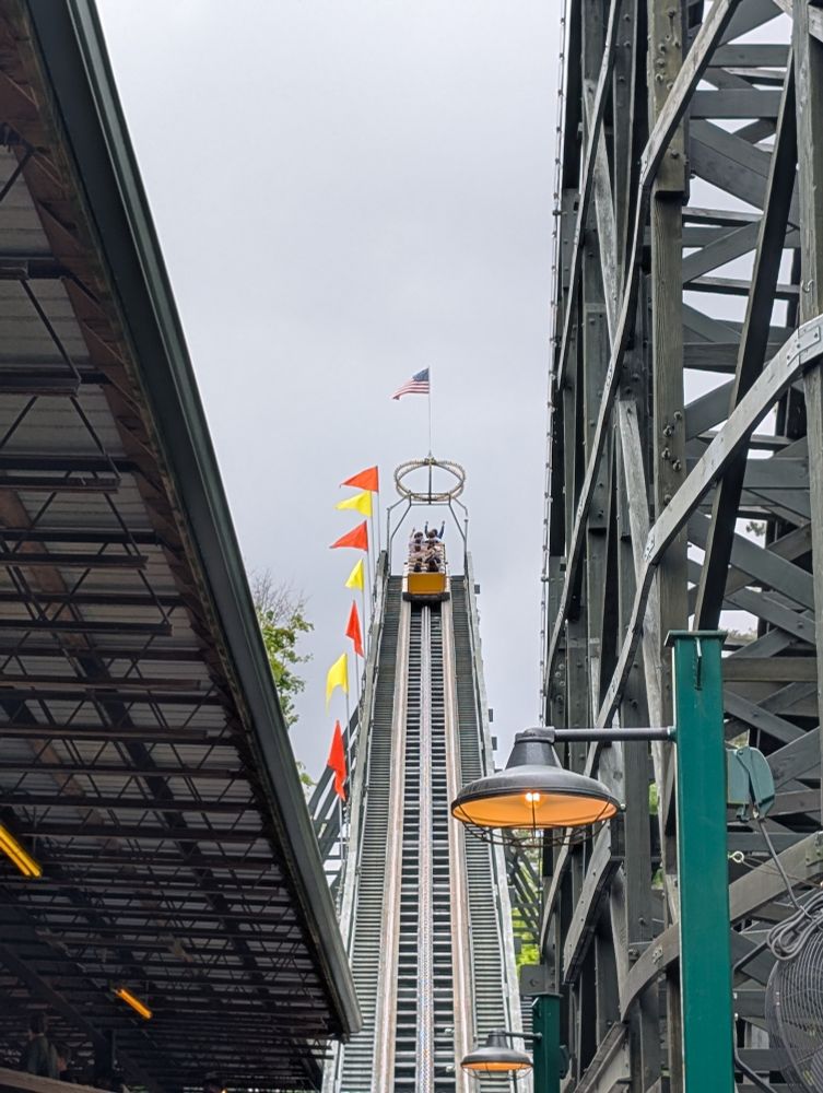 View looking up from behind the first and largest incline on a wooden roller coaster. The train is creating the top of the hill, which is dotted with yellow and orange flags