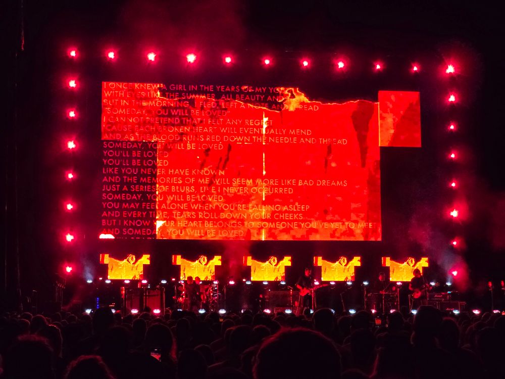 A large concert screen displaying glitched abstract graphics along with the lyrics for "Someday You Will Be Loved" as the band performs