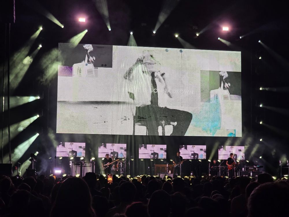 A large concert screen displaying abstract visuals while the band performs the song "Marching Bands of Manhattan"