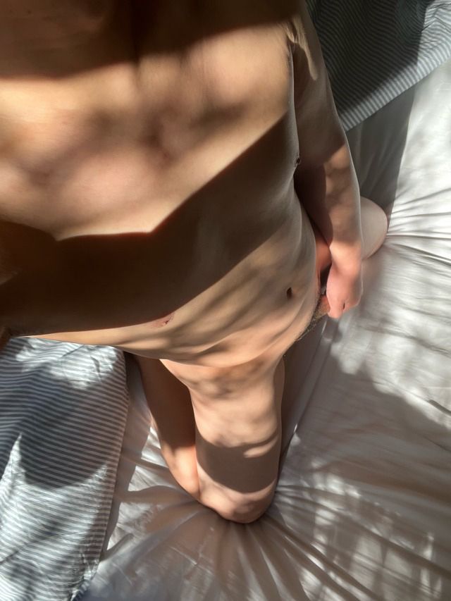 A naked man kneeling on a bed, one hand covering his crotch 