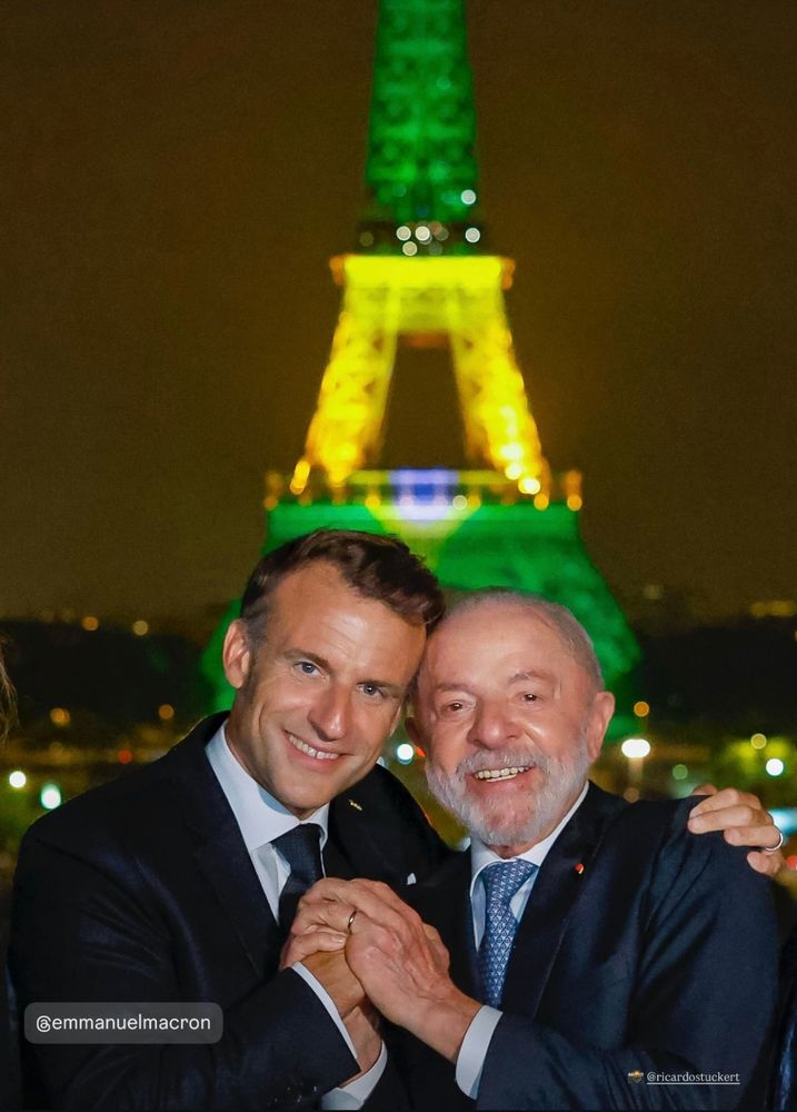 Lula e Macron de mãos dadas e cabecinhas encostadas, sorridentes, em frente à Torre Eiffel iluminada de verde e amarelo 
