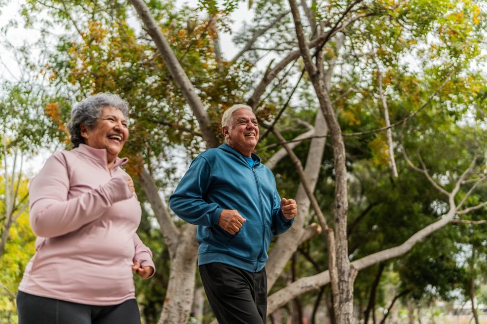 Two senior adults jogging happily in a park with trees in the background.