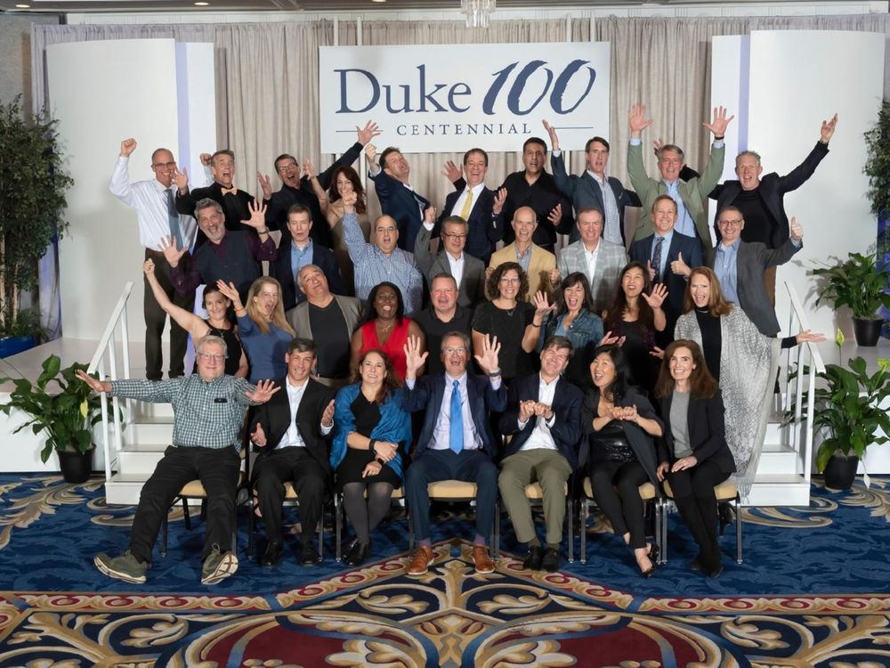 Group of people celebrating, waving with hands raised in front of a Duke 100 Centennial banner.
