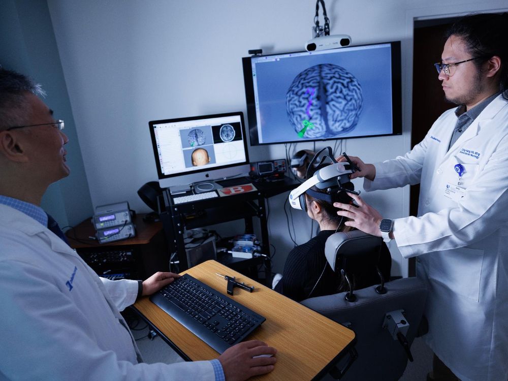 Two healthcare professionals in lab coats, one operating a computer and the other adjusting virtual reality equipment on a seated individual, in a room with multiple brain imaging screens.