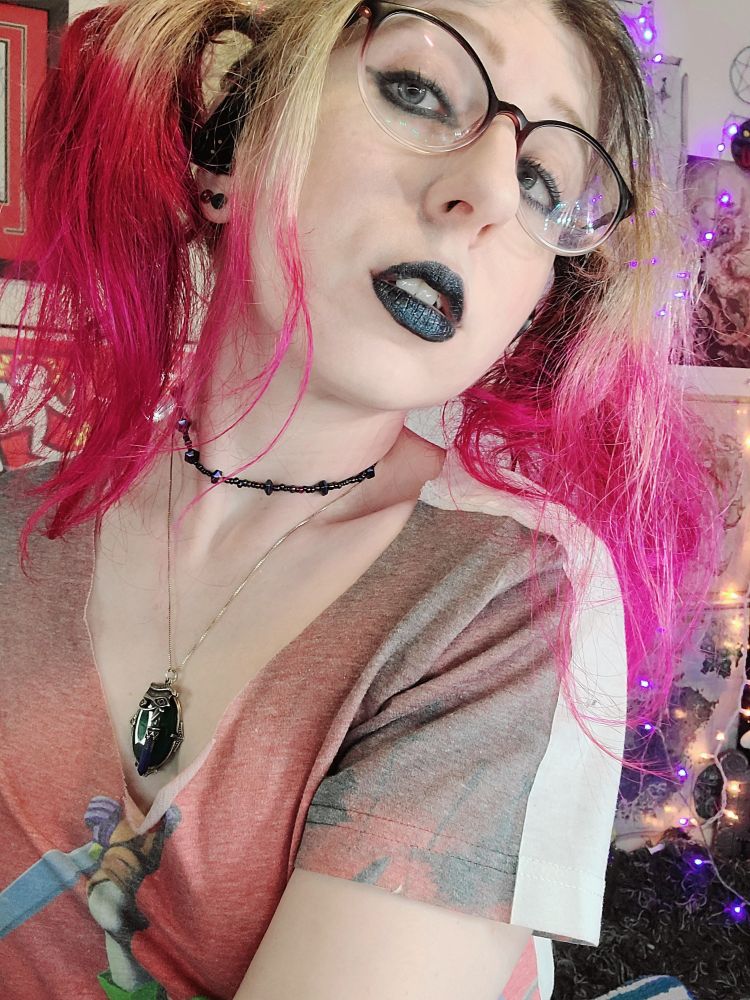 Girlbot Div staring at the viewer. They're made up in black eyeliner and lipstick with hot pink-blonde pigtails 