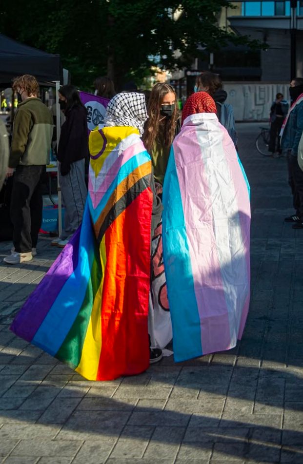 Deux manifestant·es tournent le dos à la caméra, des gens ont des pancartes en arrière plan. La personne à gauche porte un drapeau LGBTQIA2S+ la couvrant des épaules aux pieds et un keyffieh noir et blanc autour de la tête. La personne à droite porte un drapeau de la fierté transgenre la recouvrant également et peut-être un hijab (en tout cas elle a la tête couverte).
C'était la contre-manif du 21 septembre pour barrer le chemin à la mouvance transphobe autour de l'Associarion ensemble pour protéger nos enfants.
