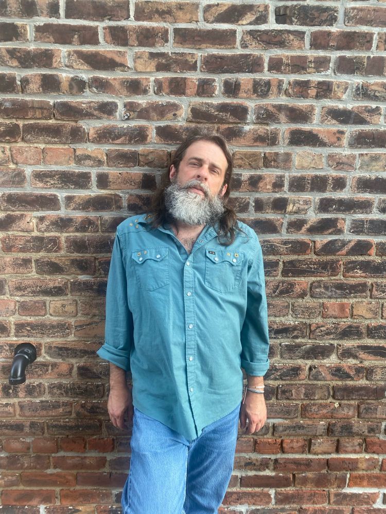 Long haired long bearded man in a western shirt and jeans stands against some Nebraska brick 