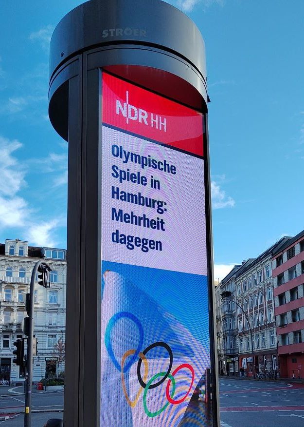 Ein digitales Display in Form einer Litfaßsäule, im urbanen Raum. Auf dem ca 3 Meter hohen Display ist eine Schlagzeile des NDR zu sehen: „Olympische Spiele in Hamburg: Mehrheit dagegen.“