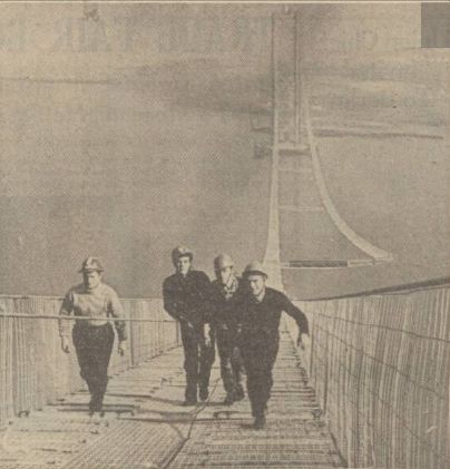 Four men in hard hats walking up a precarious-looking walkway, with a suspension bridge tower visible behind them