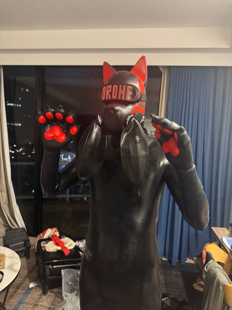 Image: a black and red silicone kitsune drone wearing a visor that reads "DRONE" in red text, it has rebreather bags attached, and its arms are raised to head height, showing off its paws.