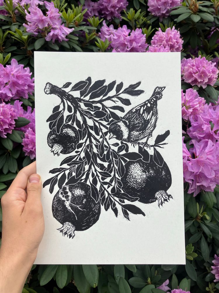 person holding a linocut print made in realistic style showing a branch of pomegranate tree. there are three fruit carved with dotted texture and an european goldfinch sitting on the branch to the right side. behind the print, there are pink rhododendron flowers 