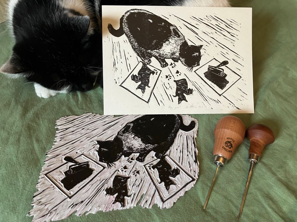 a linocut print of a curious black and white cat stepping on fresh prints and inky impressions on the floor. on the print, there is a glass slab, brayer and block to. next to the print is a sleeping cat represented on the print, the block and two carving tools 