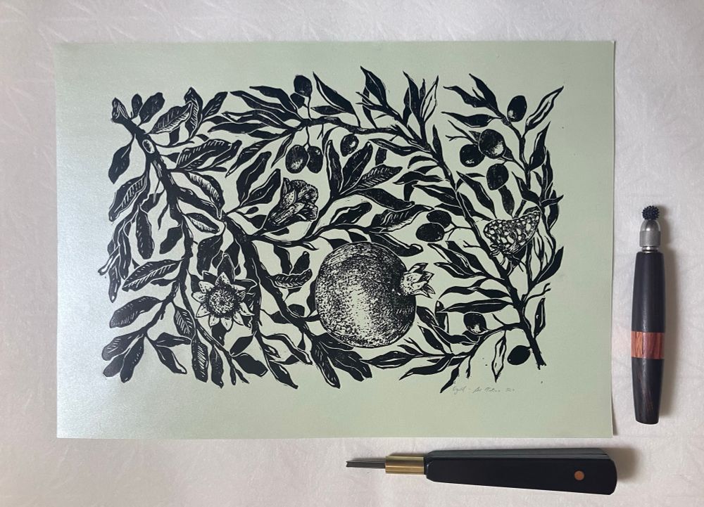 a linocut print printed with black ink on shimmery pale green paper, showing intertwined branches of pomegranate tree (with fruit and flowers) and olive tree (with fruit). there is a v gouge carving tool and relief roulette next to the print