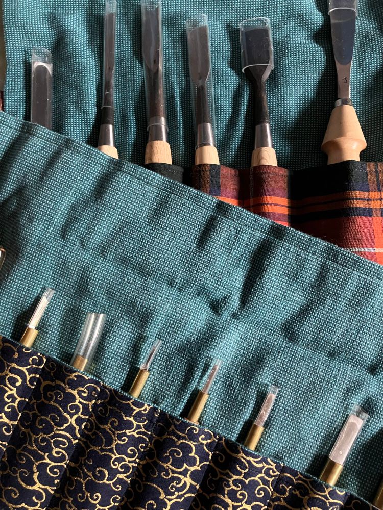 two rolls with carving tools and chisels. one is made with golden clouds pattern on navy fabric and teal canvas lining and another is made with red, orange and navy plaid pattern fabric and teal canvas lining.