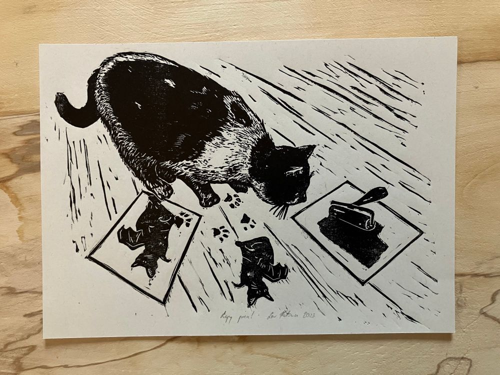 a linocut print of a curious black and white cat stepping on fresh prints and inky impressions on the floor. on the print, there is a glass slab, brayer and block.