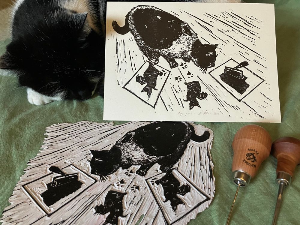 a linocut print of a curious black and white cat stepping on fresh prints and inky impressions on the floor. on the print, there is a glass slab, brayer and block. next to the print is a sleeping cat represented on the print, the block and two carving tools 