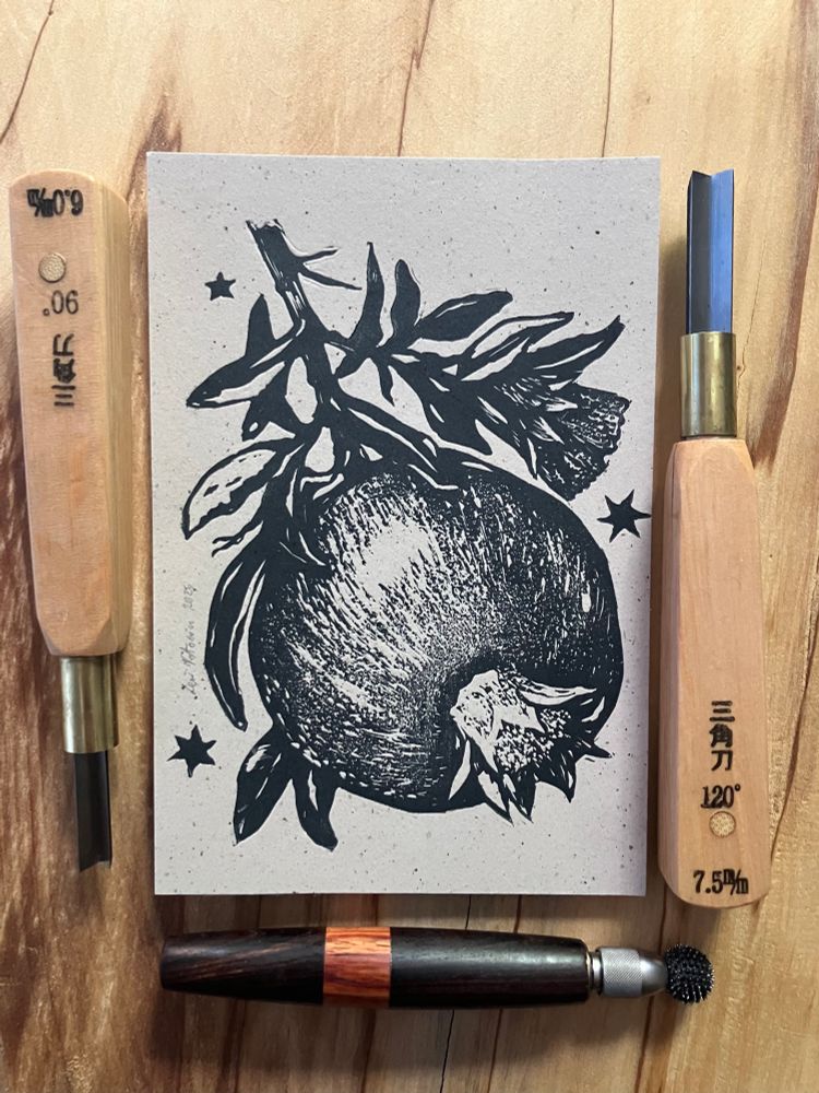 a relief print printed with dark grey ink on light brown speckled paper, representing a pomegranate branch with one fruit and one flower. there are three stars around the design. next to the print, there are two v gouges and one relief roulette.