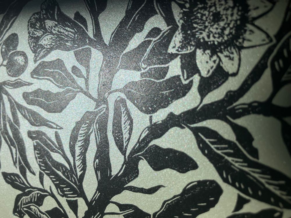 a closeup of linocut print printed on shimmery pale green paper showing a  branches, leaves and pomegranate flower 