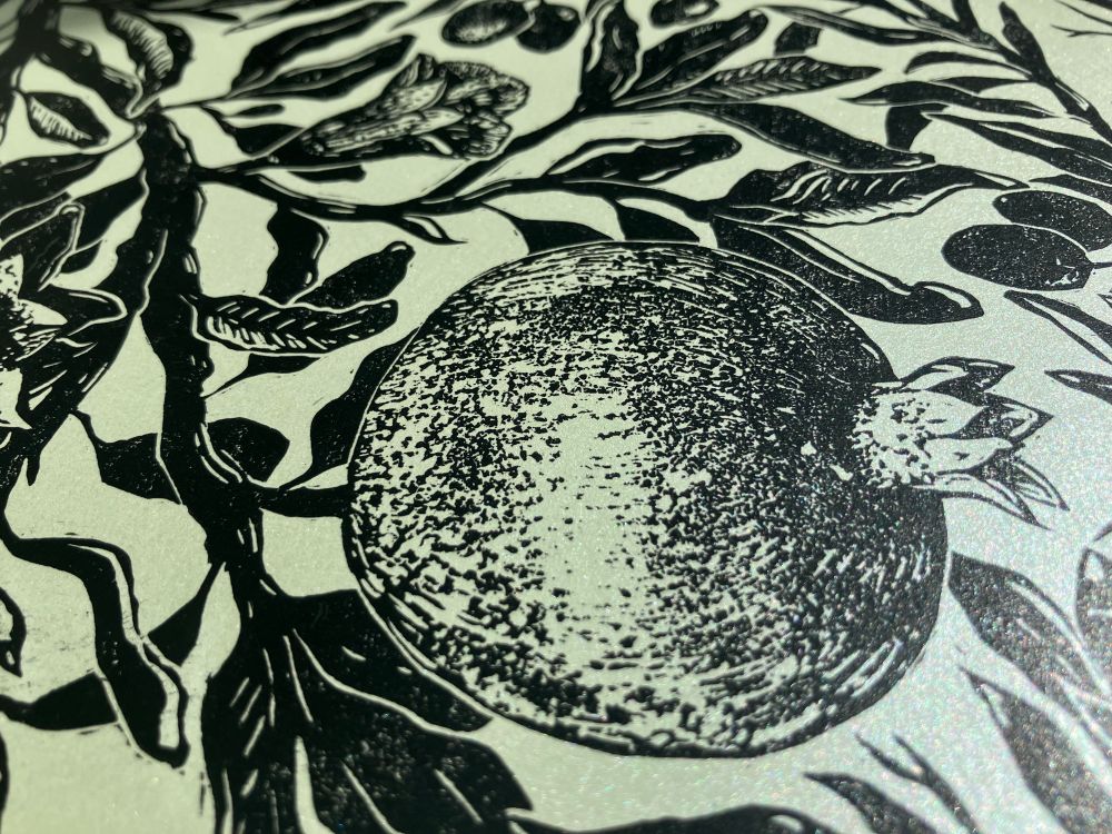 a closeup of linocut print printed on shimmery pale green paper showing a  pomegranate, made with soft roulette textures 