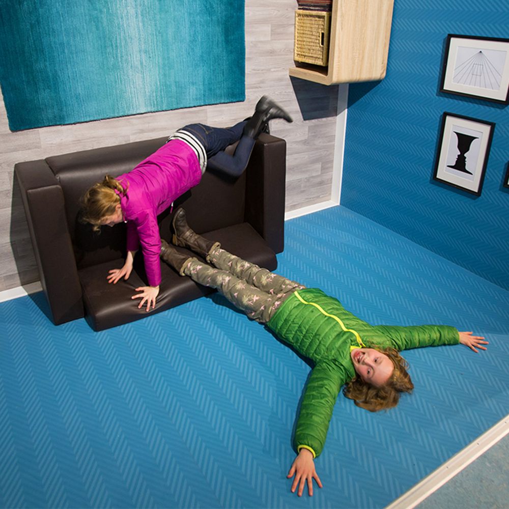 Two children appear to defy gravity in a tilted room: one in a pink jacket is on a couch, while the other in green lies on the floor.