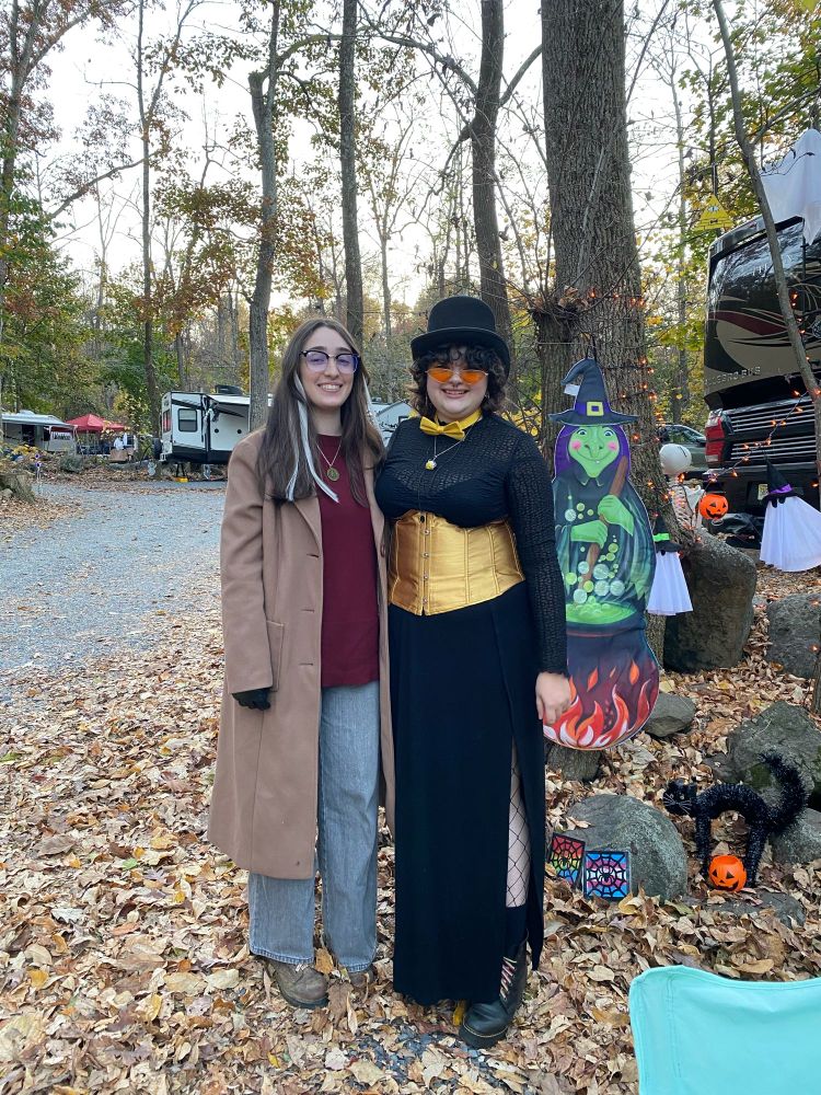 the person on the right dressed in a tan trench coat overtop a maroon shirt and grey jeans with square glasses and brown hair with grey streaks. the person on the left dressed in a black mesh shirt and black slit skirt with fishnets underneath as well as a golden yellow corset, yellow bow tie, yellow triangular glasses and a top hat